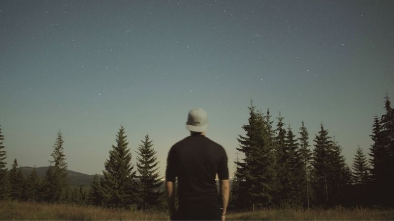 A man wearing a hat stands alone in a dark field under the night sky, surrounded by shadows and stars