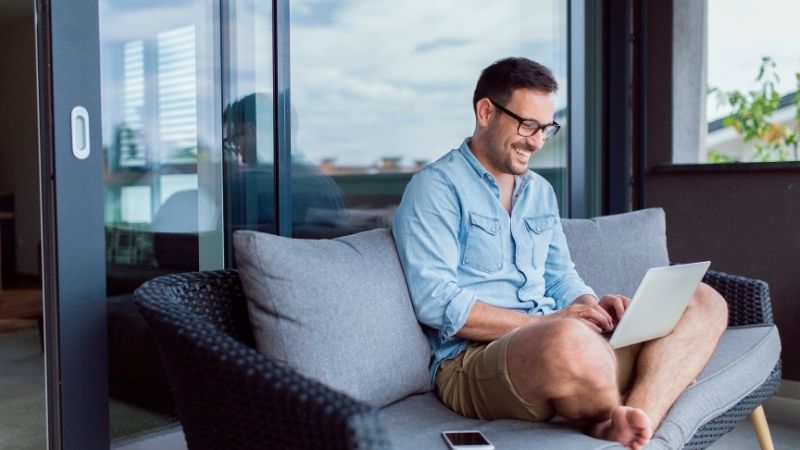 A man wearing a blue shirt working on a laptop in a modern living space with large windows, depicting remote work