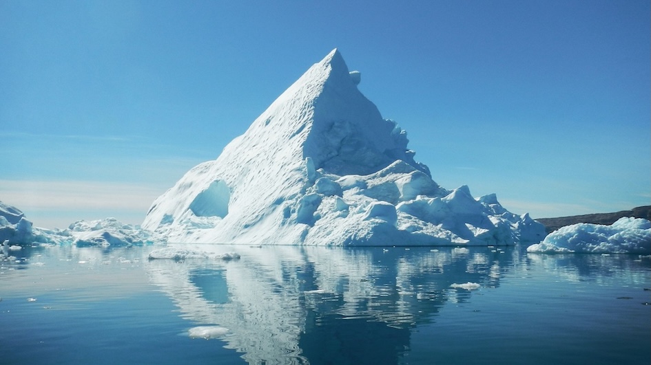 Majestic iceberg reflecting on calm sea under blue sky. The majority of the iceberg is submerged beneath the waterline, unseen.