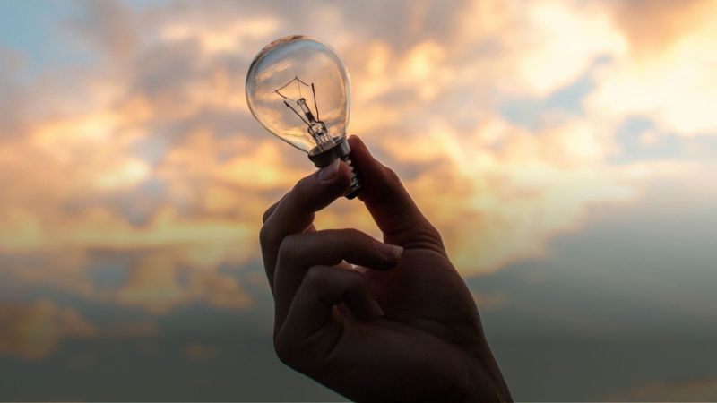 Hand holding a lightbulb against a cloudy sky at sunset