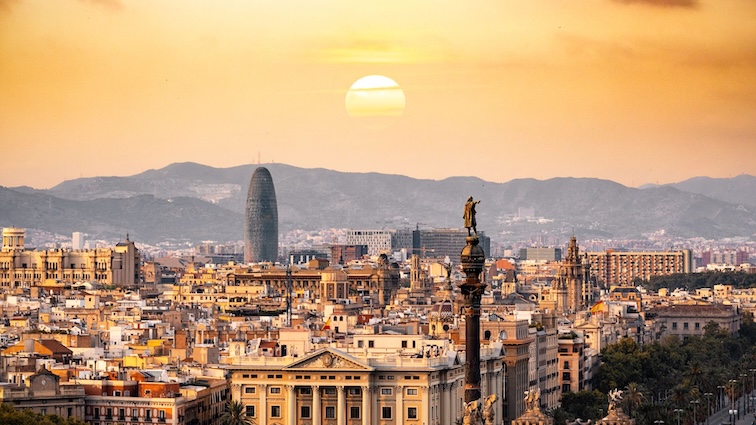 A panoramic view of Barcelona, Spain, at sunset, featuring iconic landmarks like the Sagrada Familia and Montjuïc hill