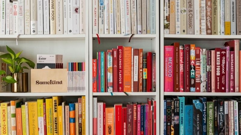 Color-coded books arranged on shelves with titles from cooking to travel, interspersed with decorative plants