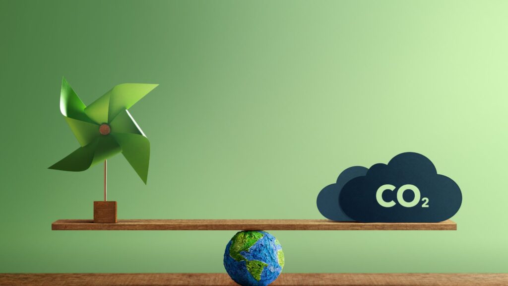 A green pinwheel and a CO2 cloud balance on a wooden scale over a small Earth model, symbolizing sustainability