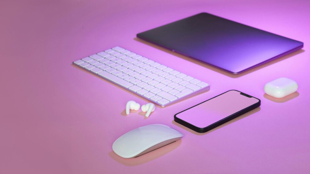 A sleek arrangement of electronic devices on a pink surface, including a laptop, keyboard, mouse, smartphone, earbuds, and charging case