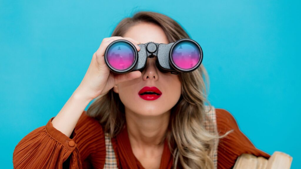 Woman with red lipstick and brown top looking through binoculars with pink-blue lenses, against a bright blue background